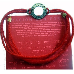 May include: Red string bracelet with a circular metal charm featuring Hebrew text. The bracelet is displayed on a red card with additional text in Hebrew and Spanish. The bracelet is a spiritual or religious item.
