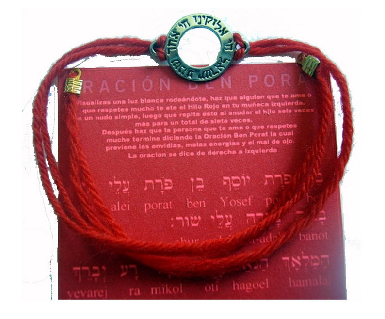 May include: Red string bracelet with a circular metal charm featuring Hebrew text. The bracelet is displayed on a red card with additional text in Hebrew and Spanish. The bracelet is a spiritual or religious item.