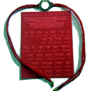 May include: A red string bracelet with a metal charm and a red card with text. The bracelet has a woven design and a metal charm with Hebrew text. The card has text in Hebrew and Spanish.