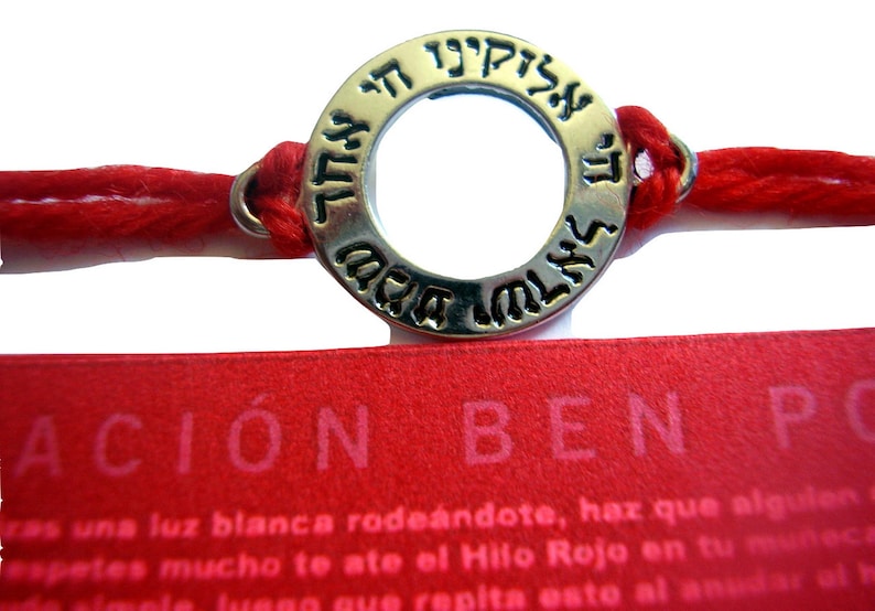 May include: Red string bracelet with a silver-colored circular charm. The charm has Hebrew text around the edge. A red tag with the word "ACION" is visible below the bracelet.