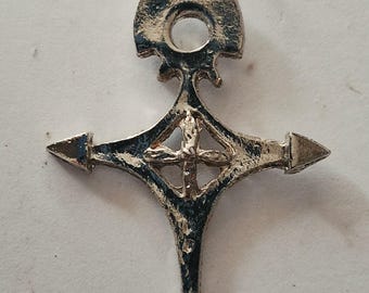 May include: A silver-toned metal pendant featuring a cross-like shape with pointed ends and a central cutout containing a smaller cross. The surface has a textured, weathered appearance with a jump ring attached at the top for hanging.