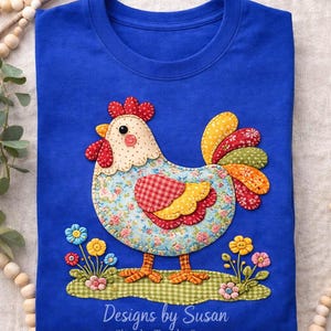 May include: A blue t-shirt featuring an appliqued chicken design. The chicken has a floral patterned body, red comb, and yellow and red tail feathers. The shirt includes the text "Designs by Susan" and "Sketchy Tees by Susan".