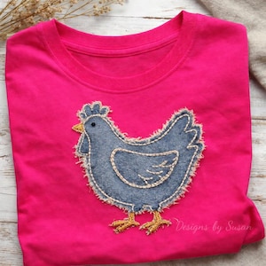 Puede incluir: Camiseta rosa brillante con un aplique de gallina de mezclilla. La gallina tiene un cuerpo azul, pico y patas amarillas, y está cosida con un borde deshilachado. El texto "Designs by Susan" está impreso en la parte inferior derecha.