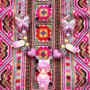 May include: A colorful necklace with a pink and white butterfly pendant, ice cream bar charms, and a pink doll figurine. The necklace is set against a vibrant, embroidered textile background with pink, red, and green floral patterns.