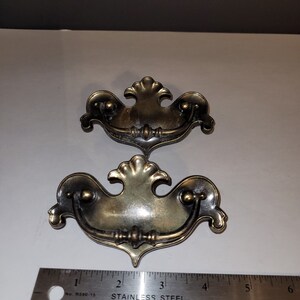 May include: Two brass-toned metal drawer pulls with ornate, scalloped backplates and swinging bail handles. A stainless steel ruler is positioned below the pulls, showing the width of each pull is approximately 5.5 inches from end to end.