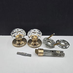 Peut inclure: Deux poignées de porte en cristal avec des bases en laiton et des composants de quincaillerie. Les boutons ont un design à facettes, reflétant la lumière. Des pièces supplémentaires incluent des plaques métalliques et un mécanisme de verrouillage.