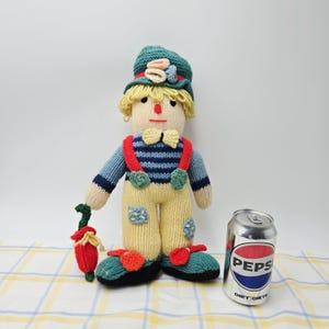May include: A hand-knitted scarecrow doll with yellow yarn hair, a teal hat, and a blue striped shirt. It wears yellow pants with patches, red suspenders, and red shoe accents. The doll holds a small red vegetable and stands beside a Pepsi soda can.