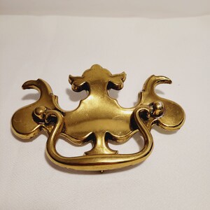 Puede incluir: Un tirador de cajón de latón pulido con una placa decorativa que presenta extensiones simétricas y curvas en forma de ala y un elemento central vertical. Un asa abatible en forma de U está unida a la placa para permitir el movimiento.