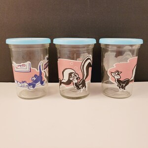 Puede incluir: Tres tarros de vidrio con tapas azules y diseños de personajes de dibujos animados. Los tarros presentan una mofeta, un gato y una mofeta y un gato juntos. Los tarros están vacíos y listos para ser llenados.