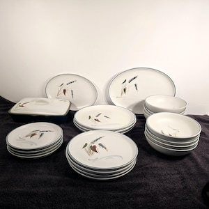 May include: A set of white ceramic dinnerware featuring a blue and green wheat design. The set includes plates, bowls, and a serving dish.