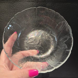 Può includere: Una ciotola di vetro trasparente con un motivo floreale dettagliato. La ciotola ha un bordo smerlato. Il design della ciotola è visibile attraverso il vetro. La ciotola è tenuta da una mano con smalto rosa.
