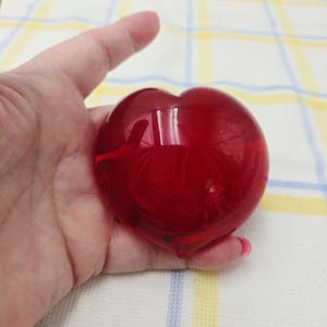 Puede incluir: Un objeto en forma de corazón de vidrio rojo intenso, liso y pulido, con una forma redondeada y simétrica. La superficie es translúcida y reflectante, captando la luz para crear brillos intensos sobre su forma curva y sólida.