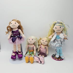 Könnte beinhalten: Vier Plüschpuppen mit lächelnden Gesichtern. Eine Puppe steht und trägt ein lila-rosa Kleid und passende Schuhe. Die anderen Puppen sitzen und tragen Kleider in verschiedenen Farben. Eine Puppe hat lange blonde Haare und ein silbernes Kleid.