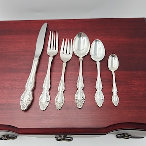 May include: A collection of silver-coloured cutlery, comprising a knife, two forks, and three spoons, displayed on a dark wooden surface. The utensils feature decorative floral patterns on their handles.