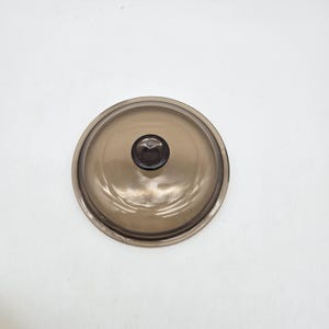 May include: A circular, transparent brown glass lid featuring a smooth, domed surface and a centred, dark brown knob handle. The lid has a slightly raised outer rim and a polished finish, designed to fit securely over a corresponding cookware vessel.