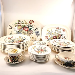 Könnte beinhalten: Ein Vintage-Geschirrset mit floralem Muster aus Porzellan, bestehend aus Tellern, Schalen, Tassen und Untertassen. Das Muster zeigt eine Vielzahl von Blumen in Rosa-, Gelb- und Blautönen auf weißem Hintergrund.