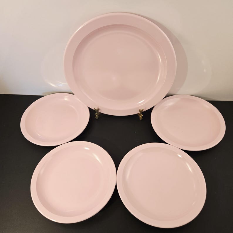 K&ouml;nnte beinhalten: Ein Set aus f&uuml;nf hellrosa Keramikplatten. Das Set beinhaltet einen gro&szlig;en Teller und vier kleinere Teller. Die Teller haben ein schlichtes, modernes Design mit leicht erh&ouml;htem Rand. Die Teller sind auf einer schwarzen Oberfl&auml;che vor wei&szlig;em Hintergrund angeordnet.