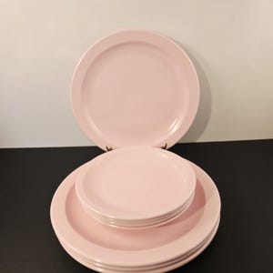 K&ouml;nnte beinhalten: Ein Satz rosa Keramikplatten, darunter gro&szlig;e Essteller und kleinere Teller, gestapelt und auf einer schwarzen Oberfl&auml;che angeordnet. Die Teller haben ein schlichtes, klassisches Design und eine glatte, gl&auml;nzende Oberfl&auml;che. Der Hintergrund ist wei&szlig;.