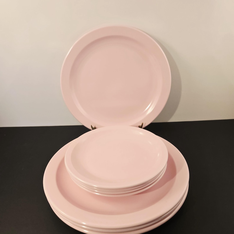 K&ouml;nnte beinhalten: Ein Satz rosa Keramikplatten, darunter gro&szlig;e Essteller und kleinere Teller, gestapelt und auf einer schwarzen Oberfl&auml;che angeordnet. Die Teller haben ein schlichtes, klassisches Design und eine glatte, gl&auml;nzende Oberfl&auml;che. Der Hintergrund ist wei&szlig;.