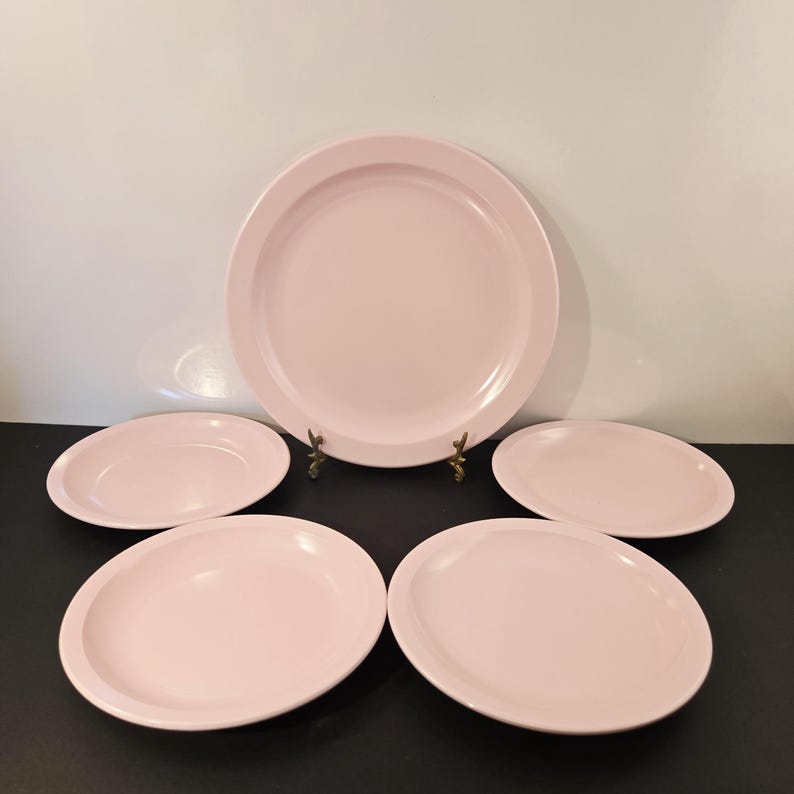 K&ouml;nnte beinhalten: Ein Set aus f&uuml;nf hellrosa Keramikplatten. Das Set umfasst eine gro&szlig;e runde Platte und vier kleinere ovale Platten. Die Teller haben ein schlichtes, klassisches Design und eine glatte, gl&auml;nzende Oberfl&auml;che. Die Teller sind auf einer schwarzen Oberfl&auml;che angeordnet.