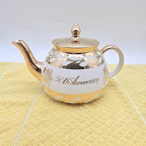 May include: A white ceramic teapot featuring a metallic gold finish on the spout, handle, lid, and base. The body is decorated with gold leaf patterns and the text 50th Anniversary in gold script lettering centred on the front of the teapot.