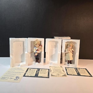 Könnte beinhalten: Zwei Porzellanfiguren von Kindern, einer ein Junge, der einen Teddybären hält, und die andere ein Mädchen, das ein Lamm hält. Beide befinden sich in ihrer Originalverpackung mit Echtheitszertifikaten. Der Junge trägt einen Hut, eine blaue Hose und braune Stiefel.