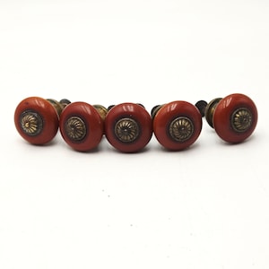 May include: A set of five round cabinet knobs featuring a smooth, deep reddish-brown finish. Each knob has a central circular brass-toned metal inlay with a detailed, radiating spiral pattern, mounted on a dark metal base and hardware stem.