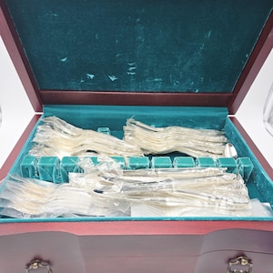 May include: A wooden cutlery chest with a burgundy exterior and a teal velvet interior. The chest is open, revealing sets of wrapped cutlery. The cutlery is silver-coloured and neatly arranged in the chest.