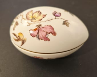 Könnte beinhalten: Cremefarbene, eiförmige Schmuckschatulle mit Deckel. Die Schatulle ist mit Blumen- und Vogelmotiven in Rosa-, Gelb- und Brauntönen verziert. Ein dünnes Goldband akzentuiert die Basis der Schatulle.