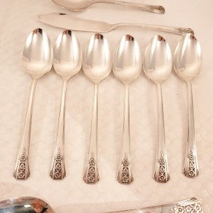 May include: Set of six silver spoons with ornate floral designs on the handles. The spoons are arranged in a row on a white surface.