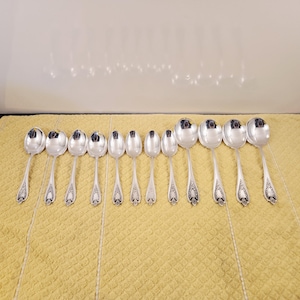 Puede incluir: Una colección de cucharas de plata dispuestas sobre una superficie amarilla texturizada. Las cucharas, de varios tamaños, presentan detalles ornamentales en los mangos. La disposición sugiere un entorno de comedor formal.