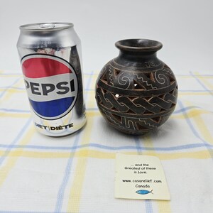 May include: A silver Pepsi Diet soda can next to a dark brown spherical clay vase with intricate cut-out geometric patterns and etched lines. A small white card features the text: ... and the Greatest of these is Love. www.casarelief.com Canada.
