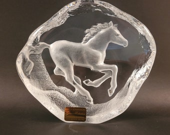 May include: A clear glass sculpture of a horse running, with a textured background. The sculpture is signed "Mats Jonasson" and "Hand made in Sweden" on a gold label.