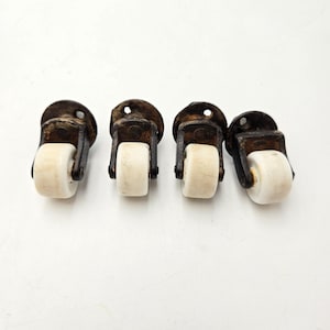 May include: Four antique furniture castors with white wheels and dark brown metal brackets. The castors are arranged in a row against a white background. Each castor has a small, round wheel and a mounting plate with a single hole.
