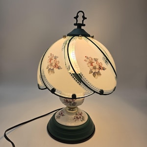 Pode incluir: Uma luminária de mesa vintage com um design floral. A luminária apresenta uma cúpula creme com flores rosa e brancas, uma base verde e um cabo preto. Os detalhes metálicos da luminária são verde escuro.