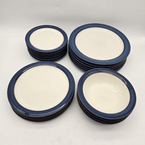 May include: A dinnerware set consisting of stacks of cream-coloured plates and bowls, each featuring a wide, dark blue rim. The collection includes multiple sizes of round plates and deep bowls, all sharing a consistent two-tone colour scheme.