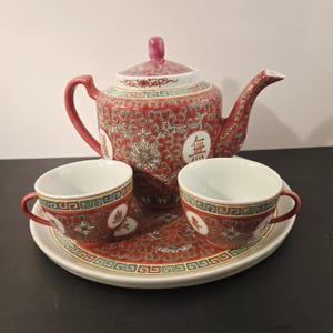 May include: A red and white porcelain tea set comprising a teapot, two teacups, and a serving tray. The set features a floral pattern and Chinese characters. The teapot has a curved handle and spout, and the teacups have small handles.