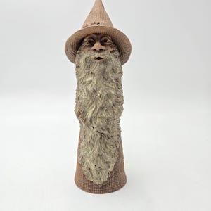 May include: A brown ceramic figurine of a bearded figure wearing a pointed hat with a textured surface. The figure features a long, flowing, light-coloured beard that extends down the front of the cylindrical body, with a small open mouth.