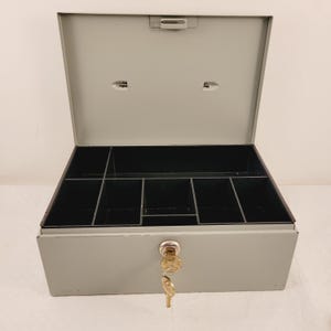 May include: A gray metal cash box with a key in the lock. The box has a compartment with multiple sections inside.