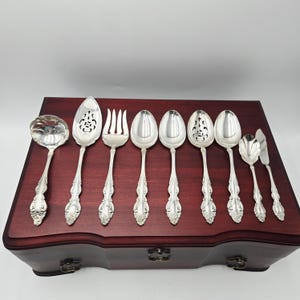 May include: An assortment of silver serving cutlery, including spoons, a fork, and a knife, displayed on a dark wooden box. The silverware has intricate floral designs and a polished finish, indicative of a formal dining set.
