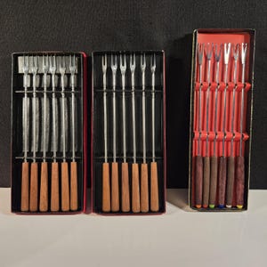 May include: Three sets of silver fondue forks with wooden handles in red boxes. The forks are arranged in rows and the handles are all the same length. The boxes are all the same size and have a red background with black trim.