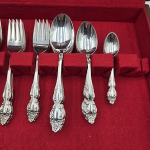 May include: A collection of decorative silverware, comprising forks, spoons, and a smaller spoon, presented in a red velvet-lined case. The silverware features an intricate floral pattern on the handles and a polished, reflective finish.