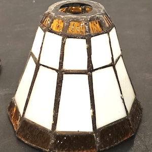 May include: A small stained glass lampshade with a geometric design. The shade features white and amber-coloured glass panels, framed by dark brown metal. The lampshade has a flared shape, with a circular opening at the top.