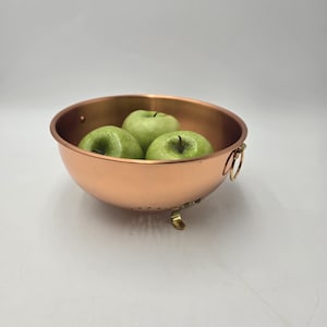 May include: A copper colander with three green apples inside. The colander has a rounded shape, small feet, and two ring handles. The apples are a vibrant green color.
