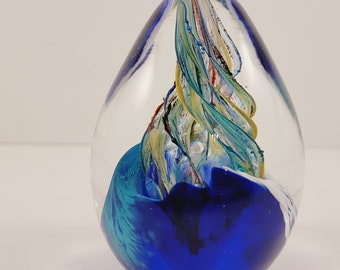 May include: A glass paperweight with a blue, white, and yellow swirl design inside. The swirl is a spiral shape and appears to be floating inside the clear glass.