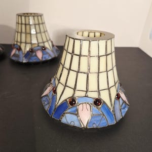Könnte beinhalten: Zwei Lampenschirme aus Buntglas. Die Schirme haben ein cremefarbenes Gittermuster und sind mit blauem, rosa und rotem Glas verziert. Die Schirme stehen auf einer schwarzen Oberfläche.