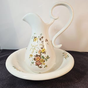 May include: White ceramic pitcher and basin set. The pitcher has a curved handle and a floral design in yellow, orange, and brown. The basin is round and also features floral accents. A classic decorative set.