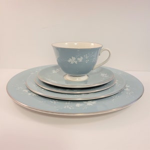 Può includere: Un servizio da tè vintage azzurro con un motivo floreale. Il set include una tazza da tè, un piattino e due piatti. I bordi sono rifiniti in argento. La tazza e i piatti hanno un motivo floreale bianco.