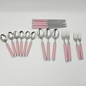 May include: A set of stainless steel flatware with pink handles. The set includes spoons, forks, and knives. The handles have a textured, ridged design. The flatware is arranged on a white surface.