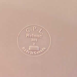 K&ouml;nnte beinhalten: Nahaufnahme einer hellrosa Oberfl&auml;che mit einem kreisf&ouml;rmigen Emblem. Das Emblem lautet "G.P.L. Melmac 303 2 Made in Canada" in einer klaren serifenlosen Schrift. Das Bild ist gut beleuchtet und der Text ist gut lesbar.